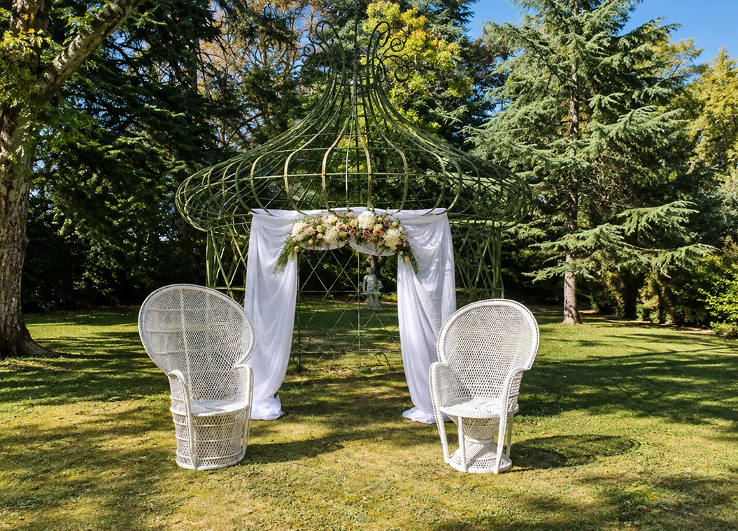 Kiosque de jardin drapé de blanc et fleuri, deux fauteuils en rotin pour la cérémonie